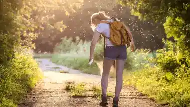 A woman with a yellow backpack stands on a sunlit path, using insect spray, to try and prevent insect bites. She's surrounded by lush greenery, conveying a peaceful outdoor setting.